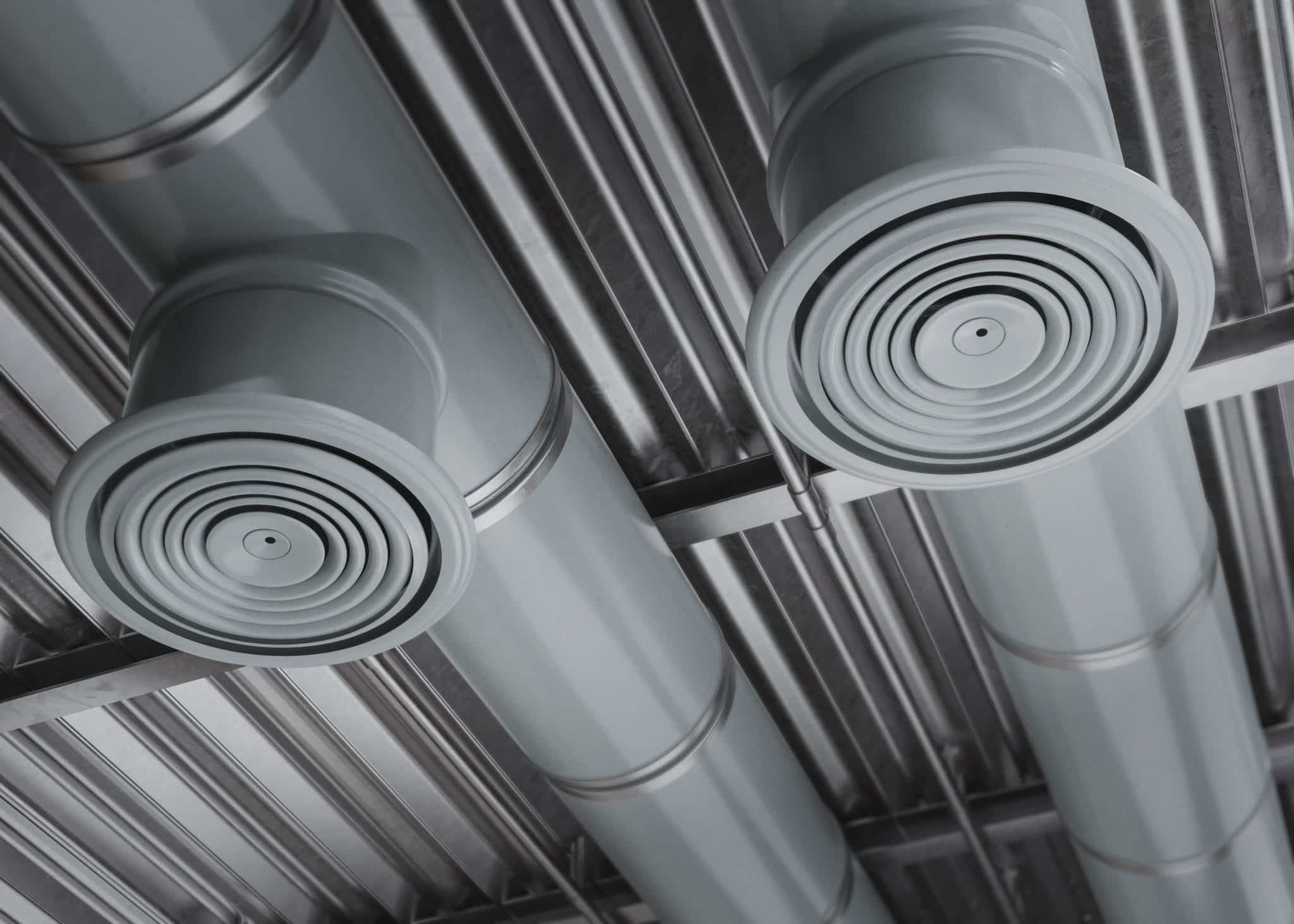 Ceiling-mounted HVAC air ducts with circular diffusers connected to exposed metal piping in an industrial space.