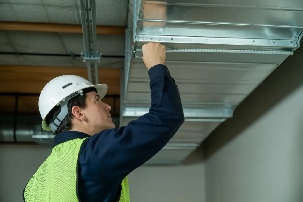 Technician wearing a hard hat and safety vest adjusting metal HVAC ductwork on an industrial ceiling